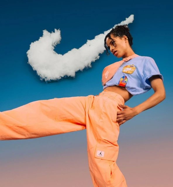 Young adult poses in bright clothes with a cloud-shaped Nike logo in a surreal promotional shot against a gradient blue background.