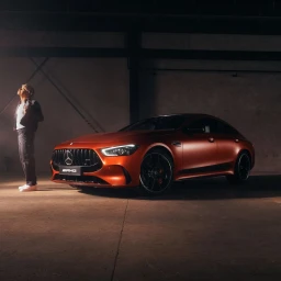 Man standing beside a glossy burnt orange Mercedes Benz luxury car in a dimly lit industrial garage interior