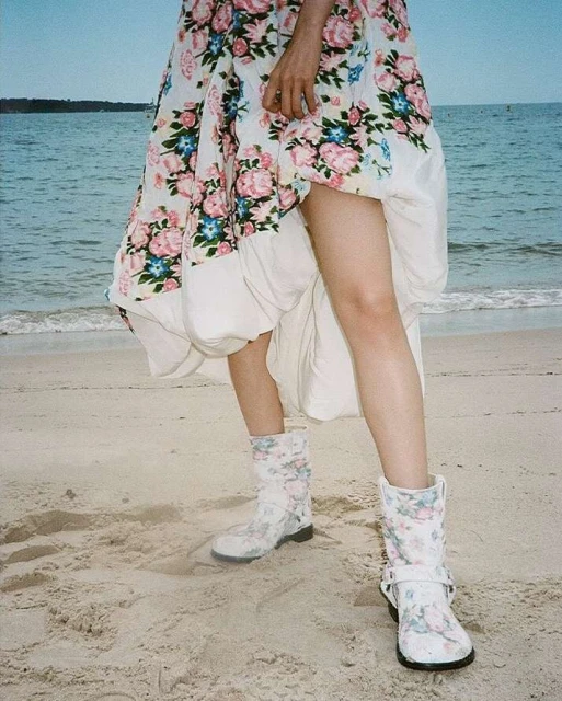 A person on a beach wearing a floral dress and floral boots poses with the ocean and sky in the background on a sunny day.