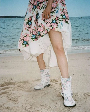 A person on a beach wearing a floral dress and floral boots poses with the ocean and sky in the background on a sunny day.
