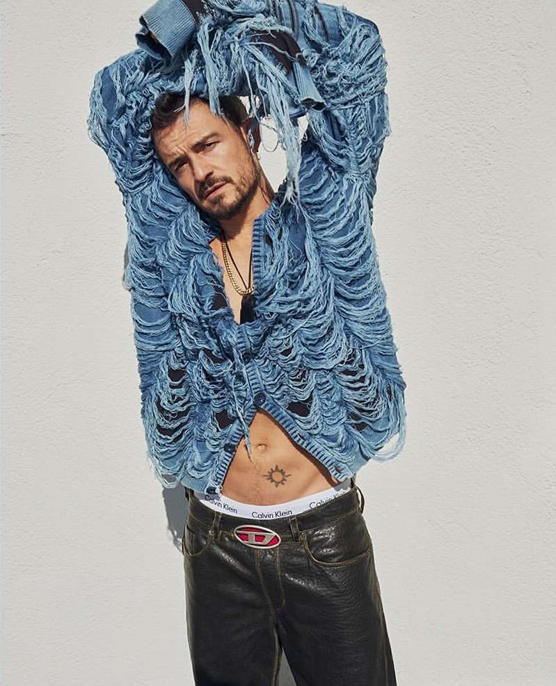 Orlando Bloom poses in a studio wearing a distressed denim jacket, black pants, and a Diesel belt, showcasing a sun tattoo on his stomach.