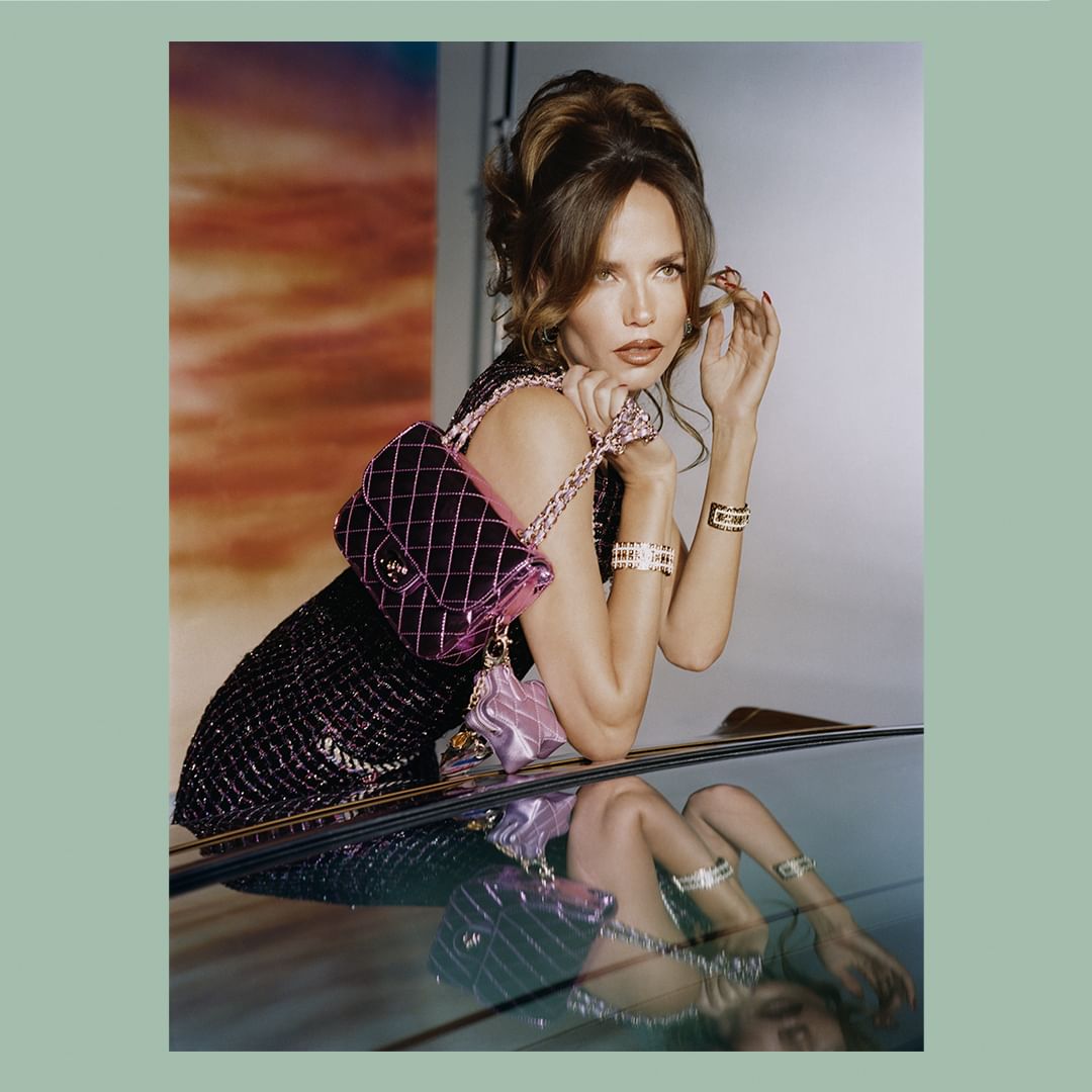Brunette woman with a vintage hairstyle poses with a black dress and pink Chanel bags, reflecting off a shiny surface in a fashion shot.
