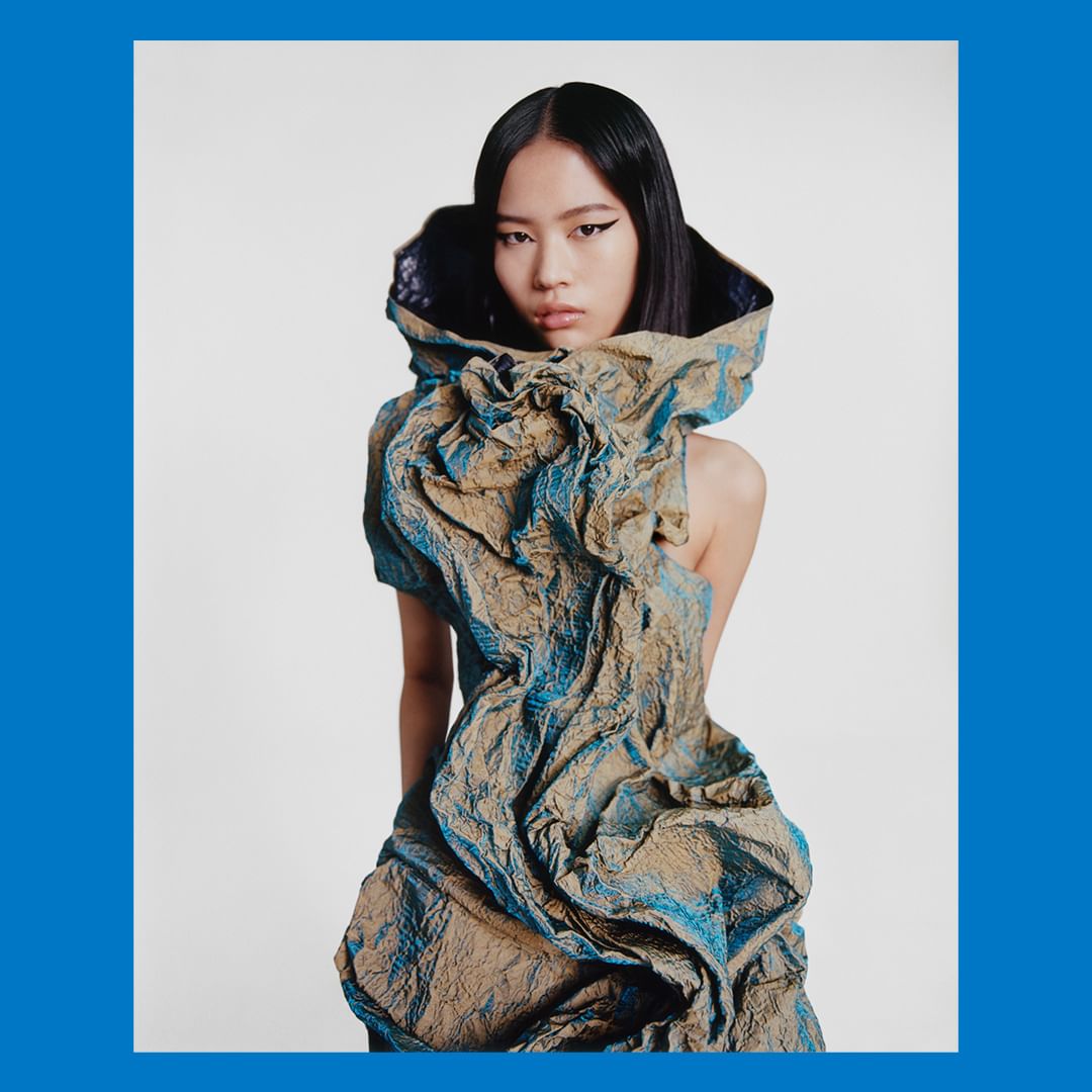 High-fashion portrait of a young Asian woman wearing an avant-garde dress with textured fabric and dramatic draping in a studio setting.