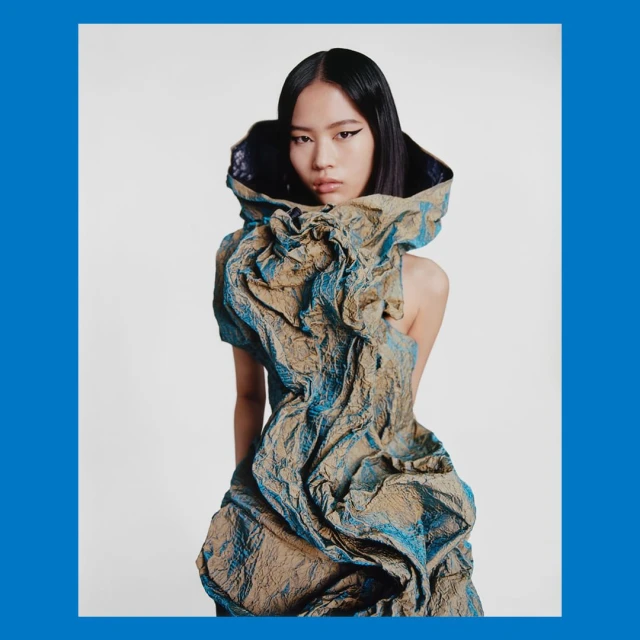 High-fashion portrait of a young Asian woman wearing an avant-garde dress with textured fabric and dramatic draping in a studio setting.