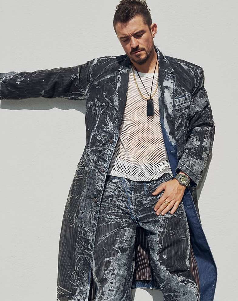 A man in a patterned coat and jeans leans against a white wall, wearing a mesh shirt, layered necklaces, and a watch in a casual outdoor portrait.