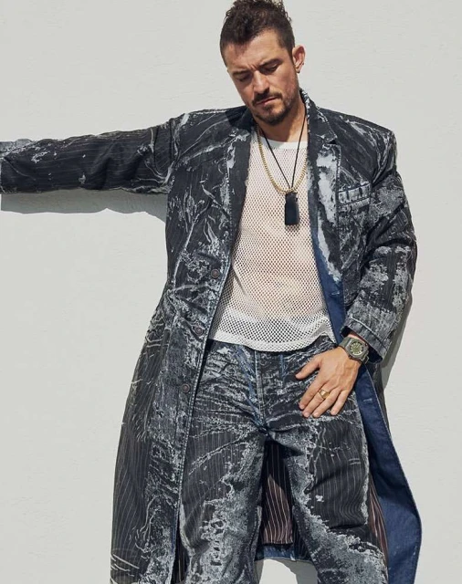 A man in a patterned coat and jeans leans against a white wall, wearing a mesh shirt, layered necklaces, and a watch in a casual outdoor portrait.