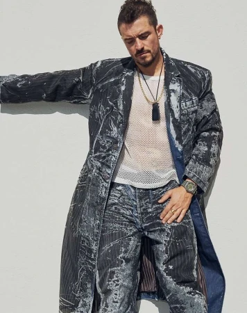 A man in a patterned coat and jeans leans against a white wall, wearing a mesh shirt, layered necklaces, and a watch in a casual outdoor portrait.