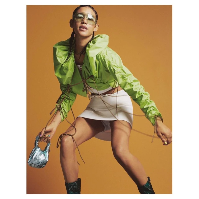 Young woman in a green jacket and white mini-skirt poses with a blue handbag in a studio setting against an orange background.