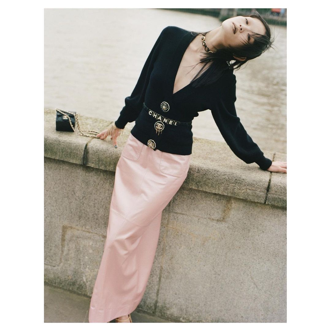 Elegant woman poses on a stone ledge outdoors wearing a black V-neck cardigan, pink maxi skirt, and Chanel belt and handbag.