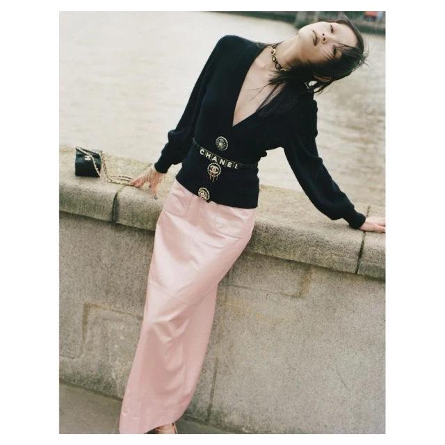 Elegant woman poses on a stone ledge outdoors wearing a black V-neck cardigan, pink maxi skirt, and Chanel belt and handbag.