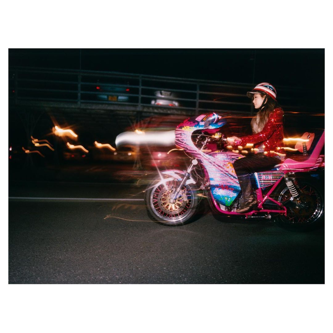 Woman in red sequin jacket and helmet riding a decorated motorcycle at night with blurred city lights creating motion effect.
