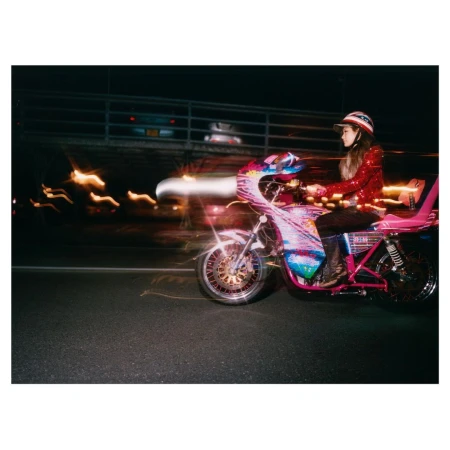 Woman in red sequin jacket and helmet riding a decorated motorcycle at night with blurred city lights creating motion effect.