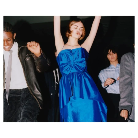 A group of young adults dressed in formal wear dance at a party, with a woman in a blue dress in the center, arms raised.