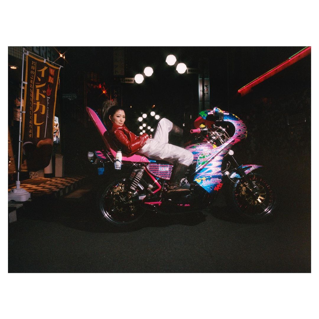 An adult female poses on a motorcycle in a nighttime outdoor urban setting, illuminated by streetlights and signage, wearing a red jacket.