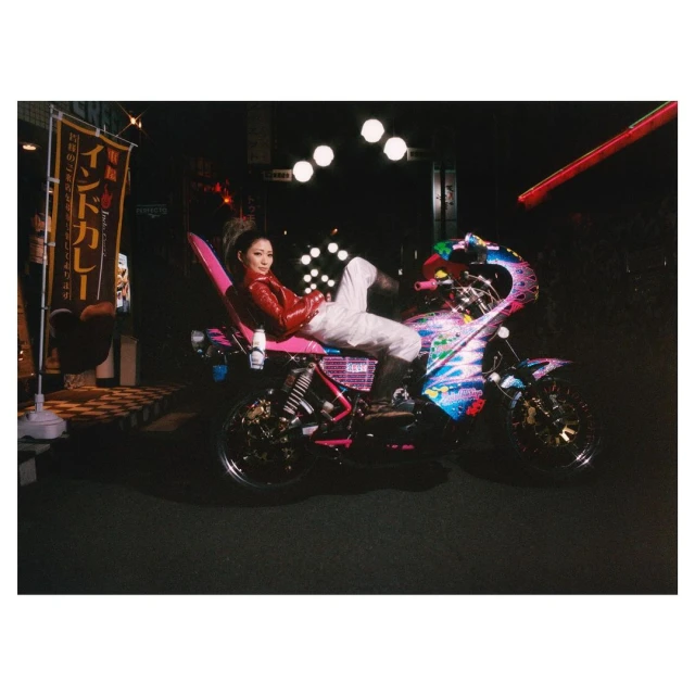 An adult female poses on a motorcycle in a nighttime outdoor urban setting, illuminated by streetlights and signage, wearing a red jacket.