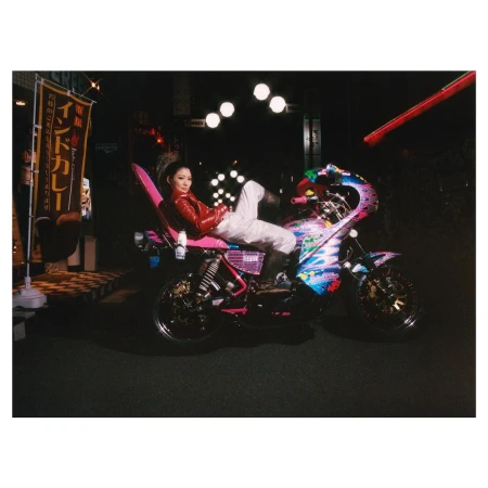 An adult female poses on a motorcycle in a nighttime outdoor urban setting, illuminated by streetlights and signage, wearing a red jacket.