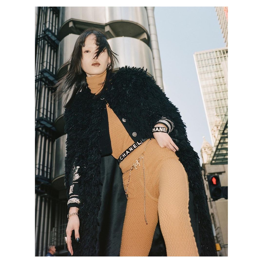 Eye-level shot of Asian woman in high fashion, wearing a textured black coat, patterned top, and trousers on an urban street.