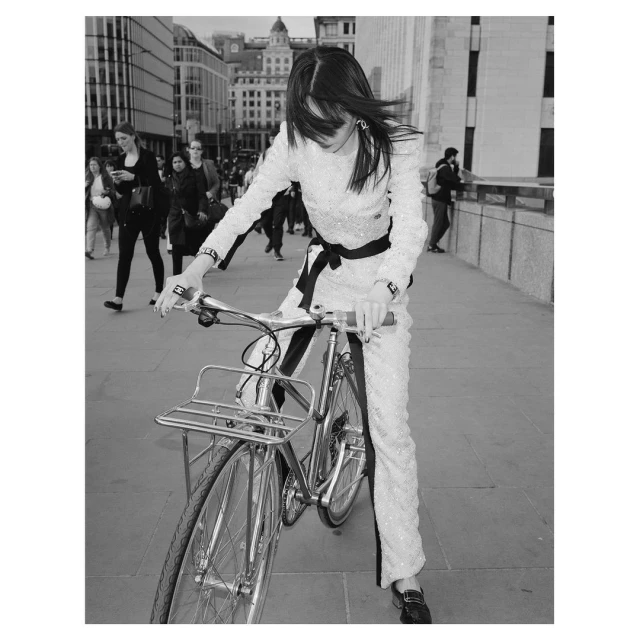 Woman in a shimmering suit poses with a vintage bicycle on a city street with buildings and pedestrians in the monochrome background.