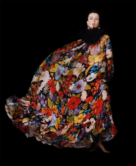 A single adult female poses in a studio wearing a long floral gown with a black feathered collar, in a fashion shot against a black backdrop.
