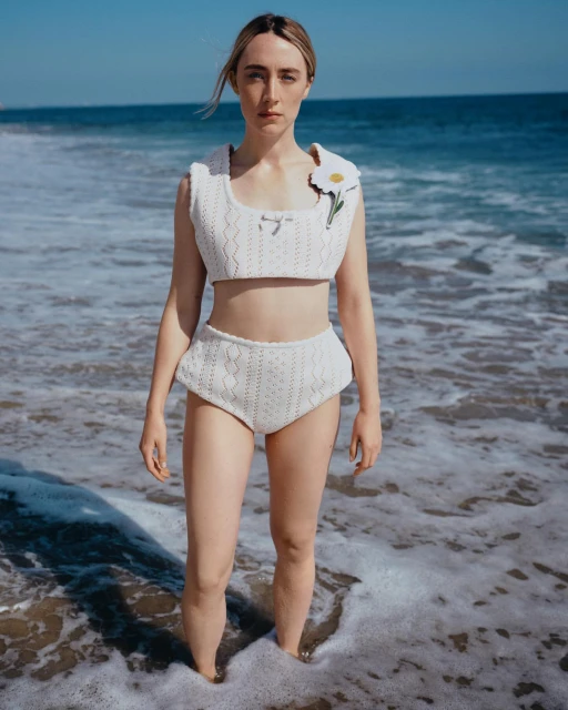 Woman in white knitted top and high-waisted bottoms stands in ocean waves on a sunny day at the beach with clear blue sky.