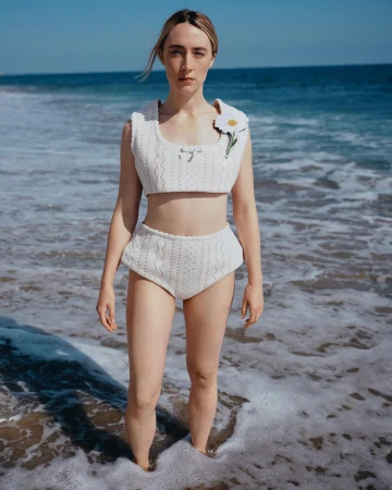 Woman in white knitted top and high-waisted bottoms stands in ocean waves on a sunny day at the beach with clear blue sky.