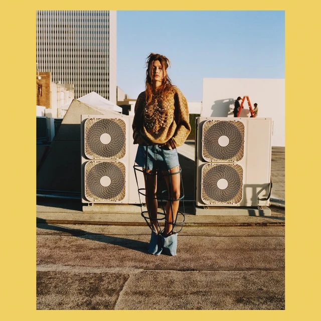 Woman poses on rooftop in sweater and denim shorts, with ventilation fans and a city building in the background on a clear day.