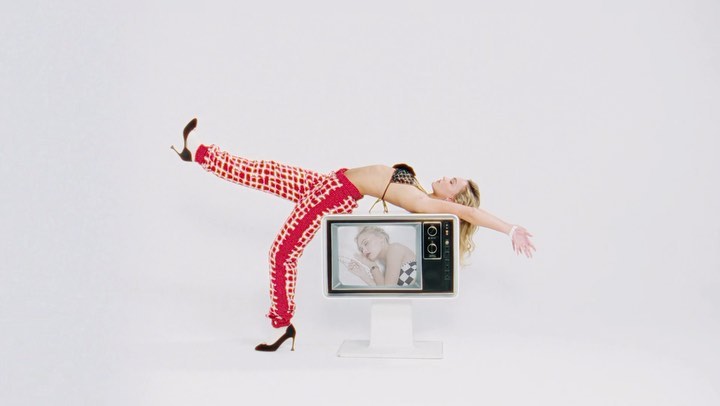 A blonde woman in a bikini top and patterned pants lies on a vintage television in a white studio, creating a playful, eye-catching composition.