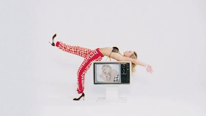 A blonde woman in a bikini top and patterned pants lies on a vintage television in a white studio, creating a playful, eye-catching composition.