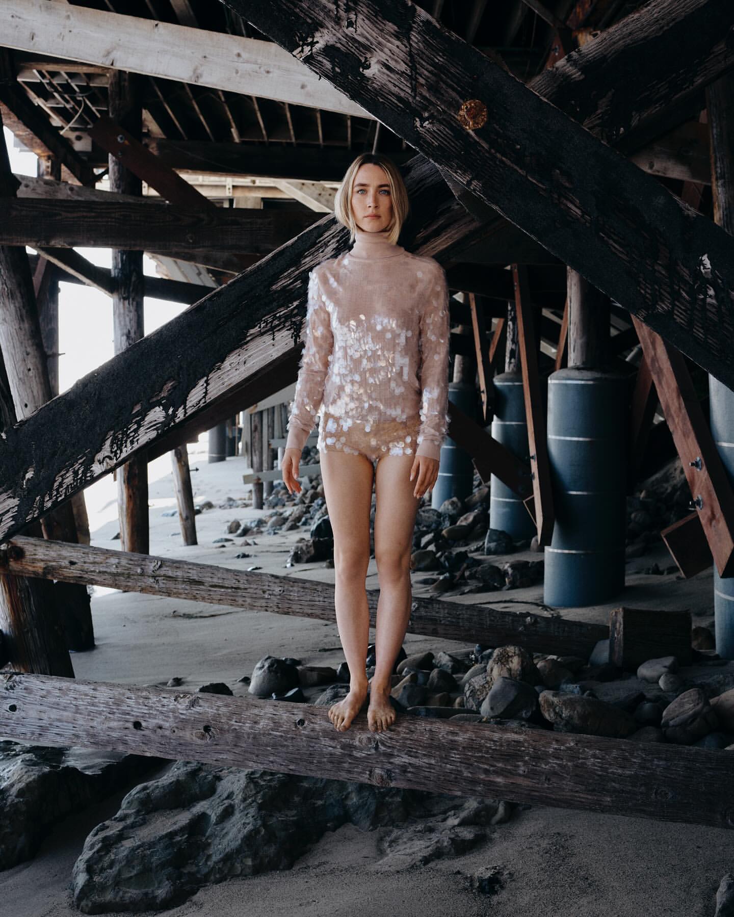 Woman in sequined top and shorts poses under wooden pier on sand beach. Natural light, neutral color palette, relaxed editorial style.