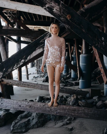 Woman in sequined top and shorts poses under wooden pier on sand beach. Natural light, neutral color palette, relaxed editorial style.