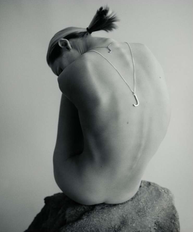 Woman with ponytail sits on a gray rock, back to the camera, wearing a silver 'J' pendant necklace in a minimalist, studio setting.