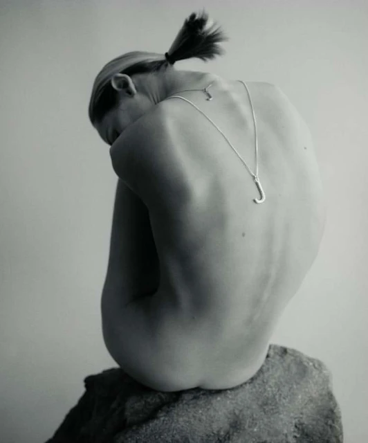 Woman with ponytail sits on a gray rock, back to the camera, wearing a silver 'J' pendant necklace in a minimalist, studio setting.