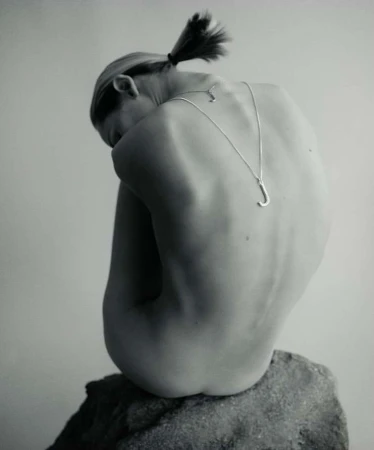 Woman with ponytail sits on a gray rock, back to the camera, wearing a silver 'J' pendant necklace in a minimalist, studio setting.