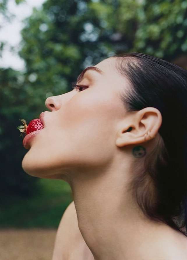 Portrait of a brunette adult female with a strawberry in her mouth outside with green foliage in the blurred background in soft lighting.