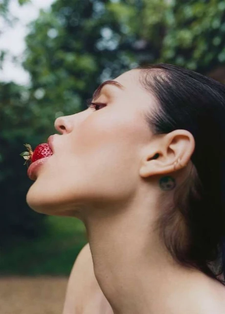 Portrait of a brunette adult female with a strawberry in her mouth outside with green foliage in the blurred background in soft lighting.