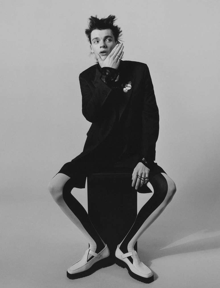 Young man in black blazer and shorts with graphic leggings and loafers strikes a pose with a hand to his cheek in a studio setting.