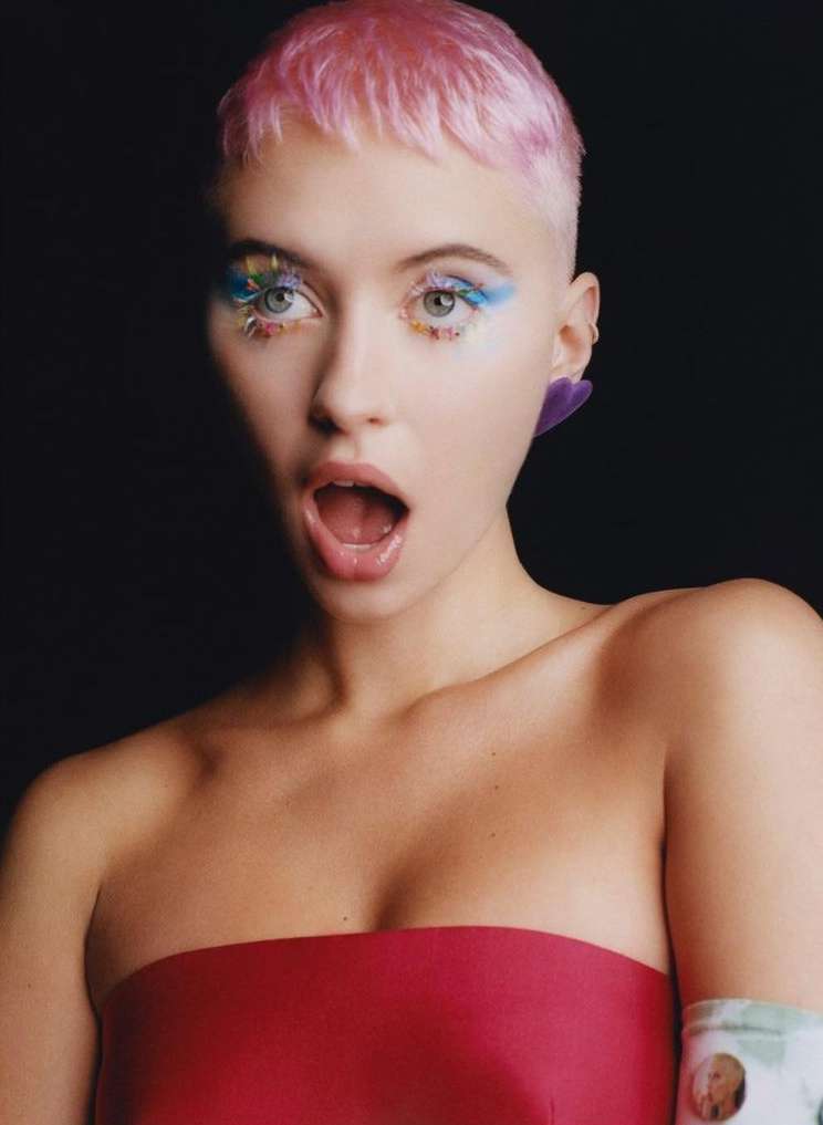 Close-up of a young woman with pink hair and creative blue eye makeup, wearing a strapless red top against a dark background.