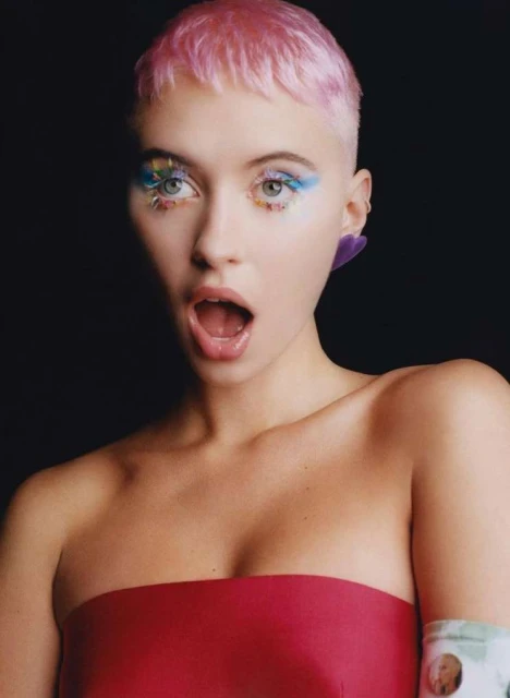 Close-up of a young woman with pink hair and creative blue eye makeup, wearing a strapless red top against a dark background.