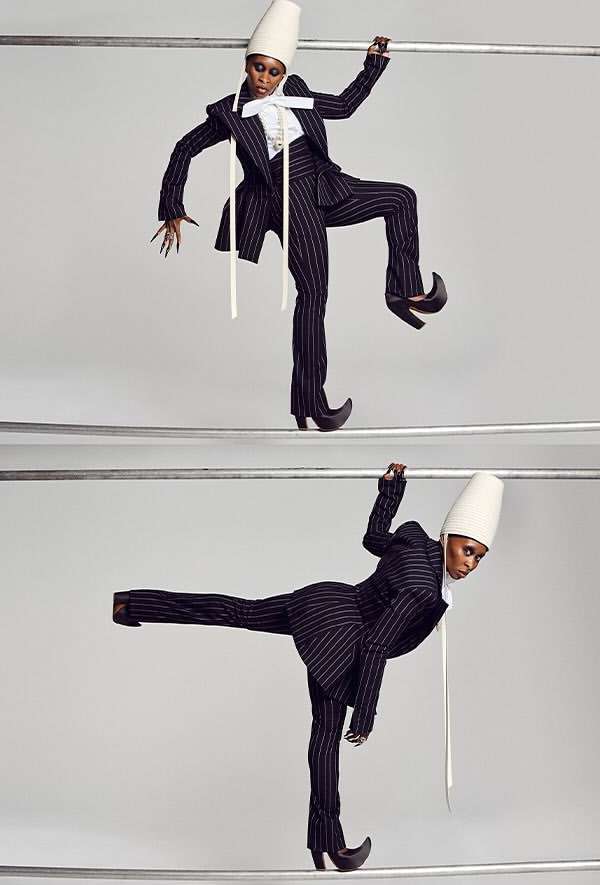 An African American fashion model balancing on rails wearing a pinstripe suit, large white bow, white cone hat, and curled-toe boots in studio.