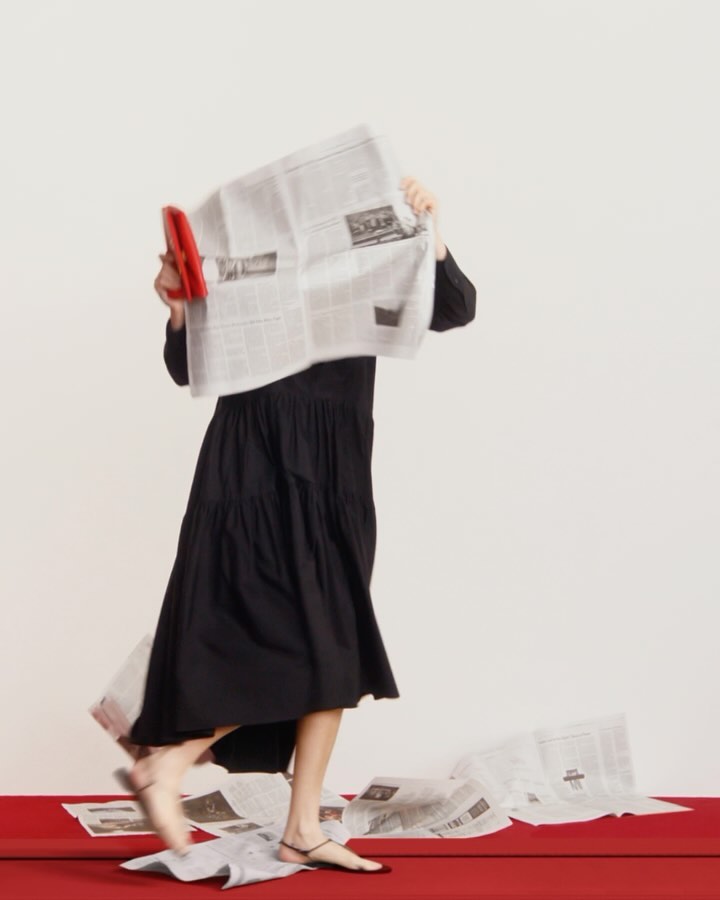 A woman in a black dress walks with a red clutch, partially obscured by newspaper against a cream background with a red floor.