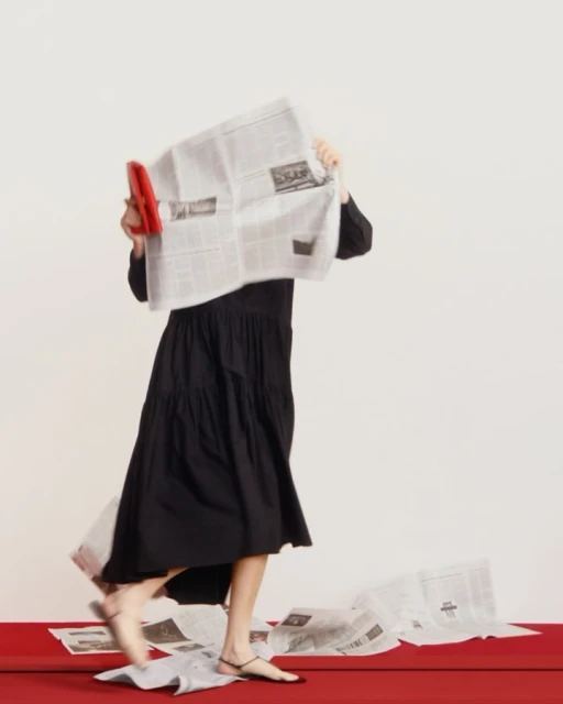 A woman in a black dress walks with a red clutch, partially obscured by newspaper against a cream background with a red floor.