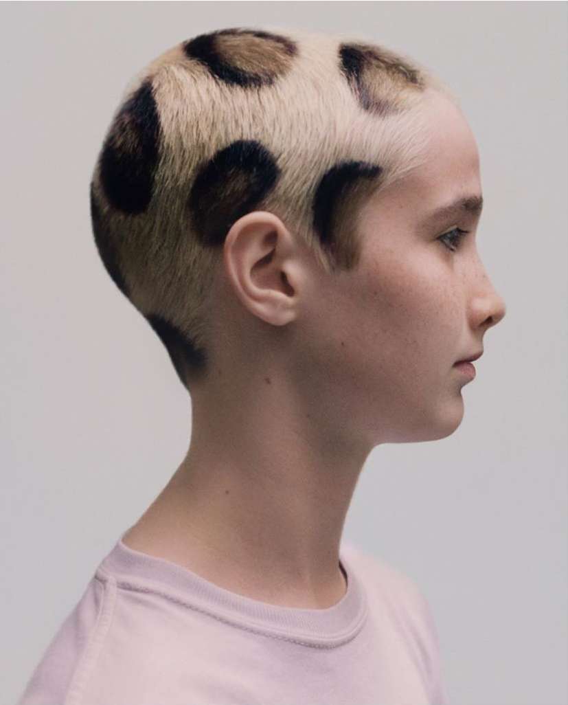 Profile of an adult female with short blonde hair featuring dark brown circular patterns, wearing a light pink t-shirt against a white background.