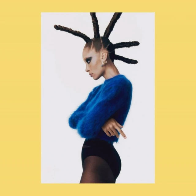 Profile of woman with creative updo, wearing a blue sweater and black bottoms, shot against a bright white backdrop.