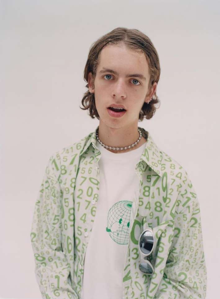 Young man in a white t-shirt and a green-and-white shirt with numbers, wearing a beaded necklace, and sunglasses in the pocket.
