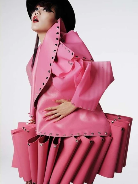 A stylish woman poses in a pink avant-garde fashion outfit with a black hat against a clean background in a studio shot.