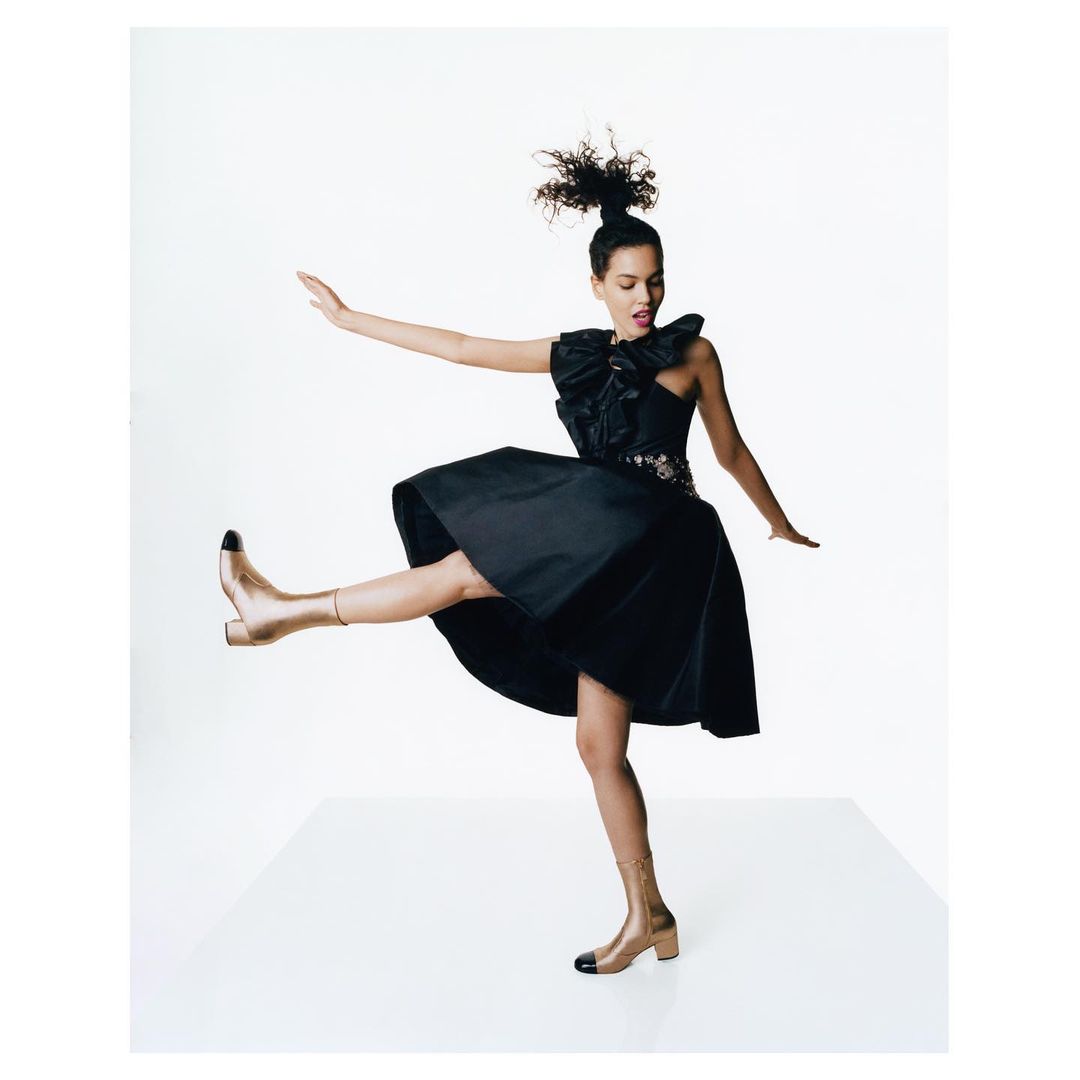 A young, stylish woman in a black dress and gold boots strikes a dynamic pose against a stark white backdrop, fashion and beauty.