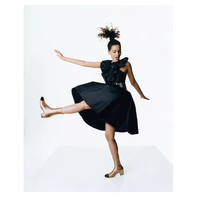 A young, stylish woman in a black dress and gold boots strikes a dynamic pose against a stark white backdrop, fashion and beauty.