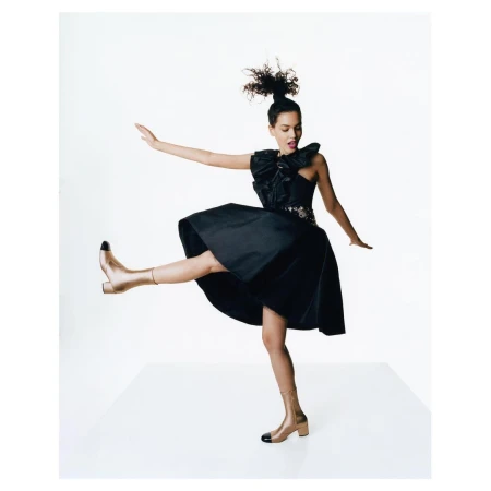A young, stylish woman in a black dress and gold boots strikes a dynamic pose against a stark white backdrop, fashion and beauty.