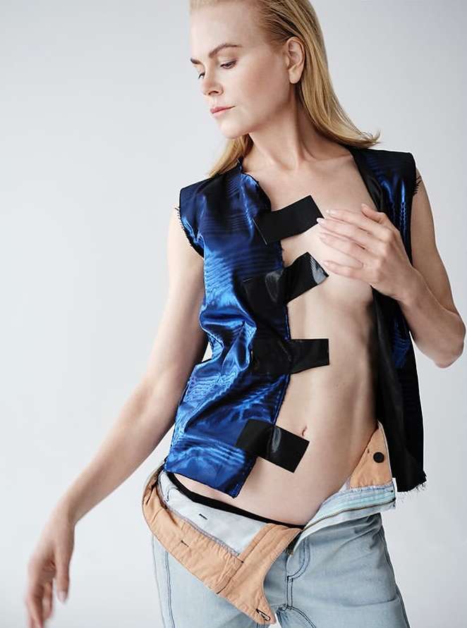 Fashion shot of adult female model, Nicole Kidman, in a blue vest with black tape, jeans slightly unbuttoned, posing against white background.