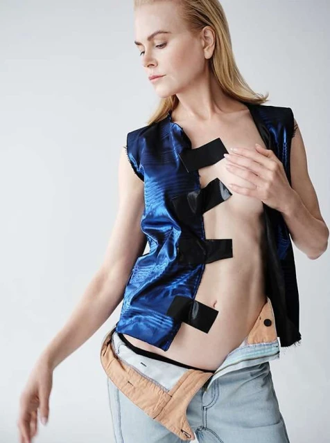 Fashion shot of adult female model, Nicole Kidman, in a blue vest with black tape, jeans slightly unbuttoned, posing against white background.